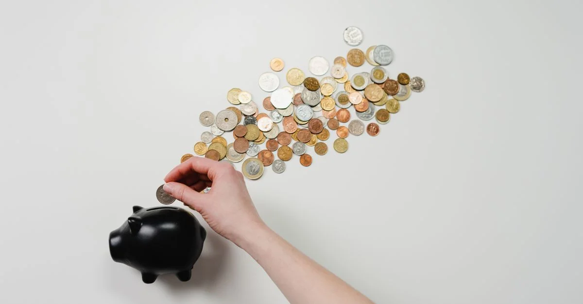 Personne plaçant une pièce dans une tirelire noire avec des pièces éparpillées, symbolisant l'épargne et la finance.