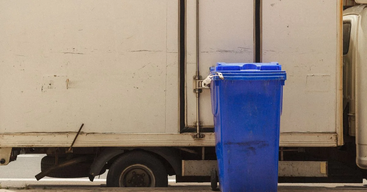 Un bac de recyclage bleu se tient à côté d'un camion de livraison garé dans une zone urbaine.