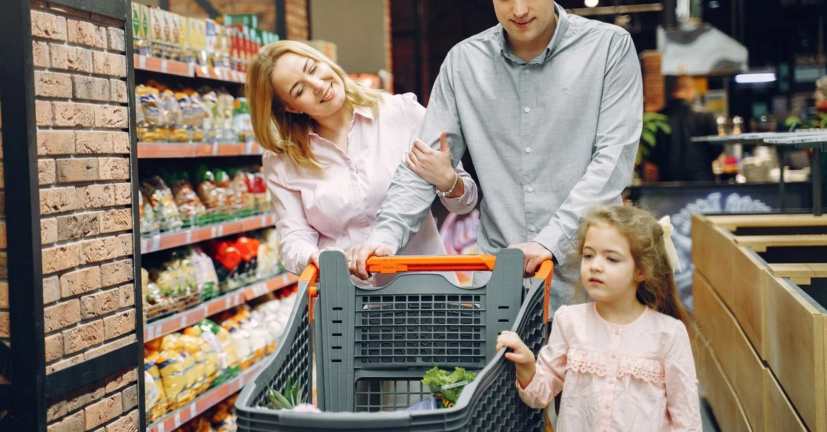 Une famille de trois personnes faisant des courses dans un supermarché, poussant un chariot plein de courses.